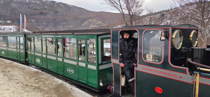 Vintage green train with a bundled conductor standing in the open doorway against a snowy mountain backdrop.
