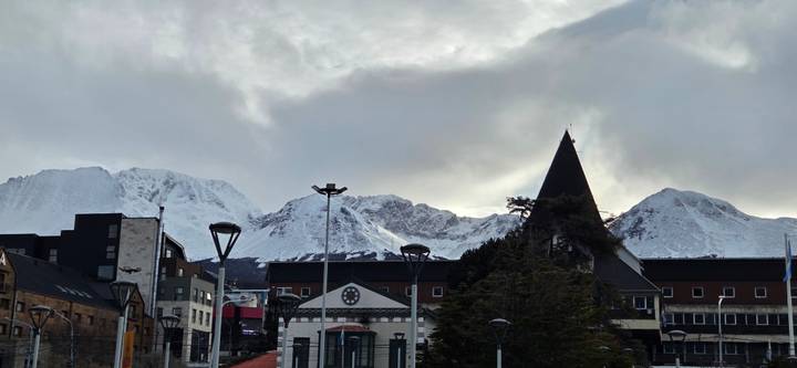 Snow-capped mountains towering over a small coastal city under a moody cloudy sky.