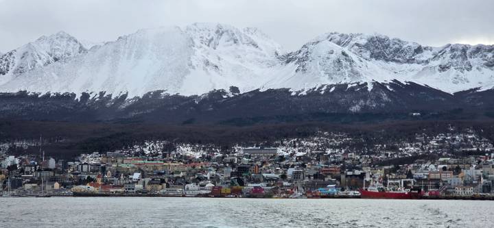 Panoramic view of Ushuaia town backed by dramatic snowy mountains, seen from the water.