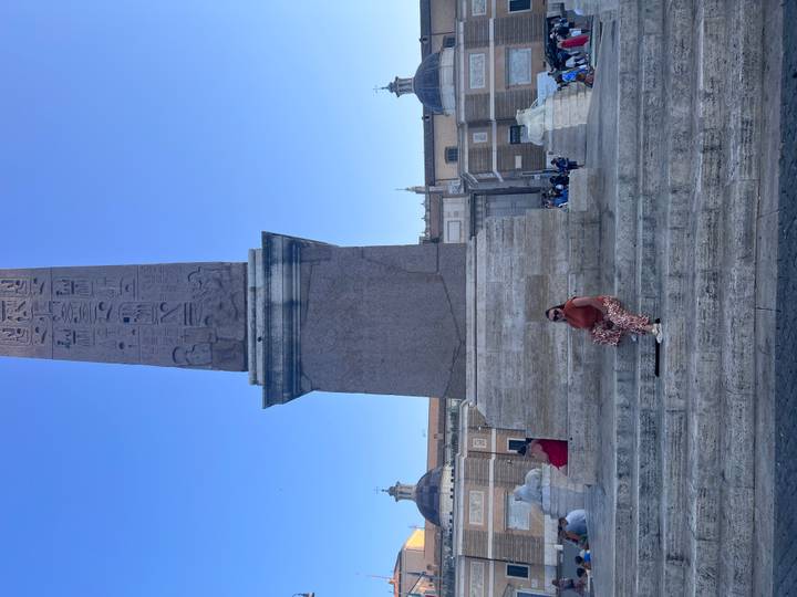 Single traveler seated on vast stone steps beneath tall obelisk and blue sky