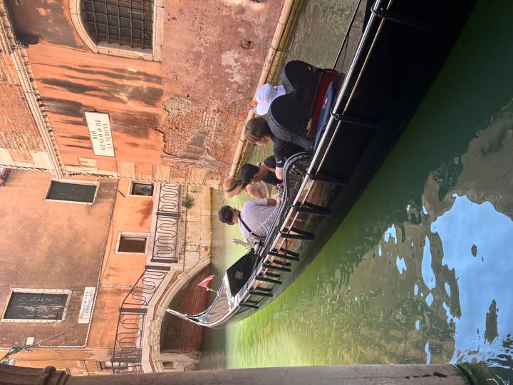A gondolier steers a family-filled black gondola along a narrow emerald canal beside Venice’s brick walls and arched bridge.
