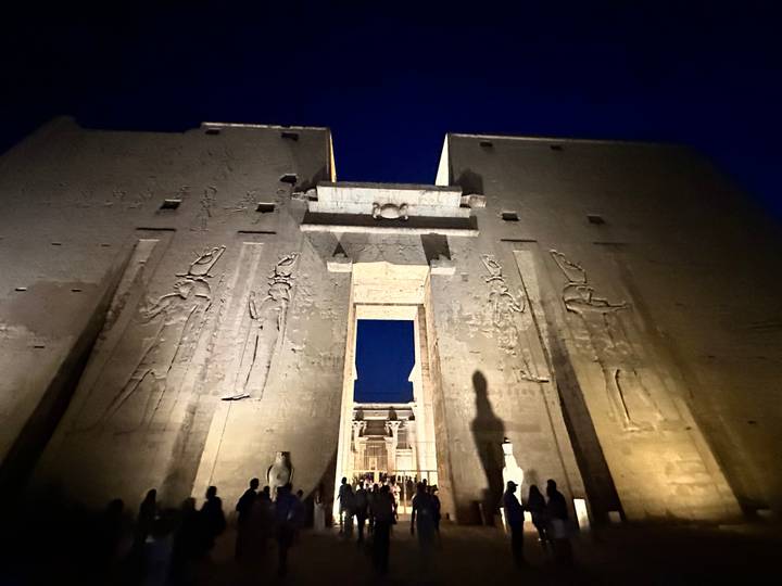 Night view of a massive Egyptian temple façade illuminated from below with crowds entering through the central gate.