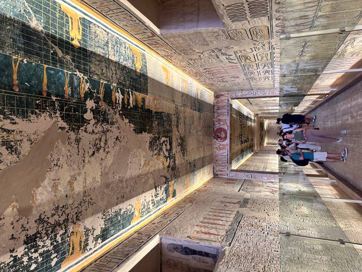 Long decorated corridor of a tomb covered in colorful hieroglyphs with visitors walking through behind glass barriers.