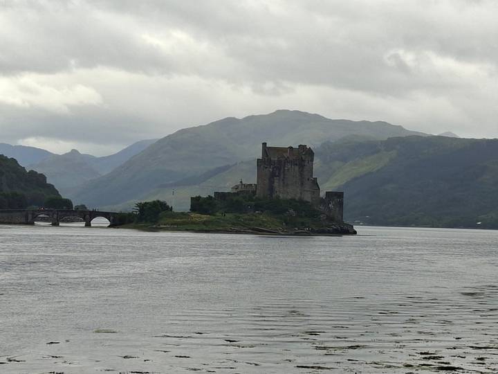 Stone Eilean Donan-like castle on an islet with arched bridge and misty hills behind