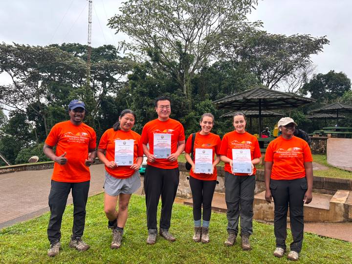 Hikers wearing matching orange shirts proudly display Kilimanjaro certificates at a lush park viewpoint