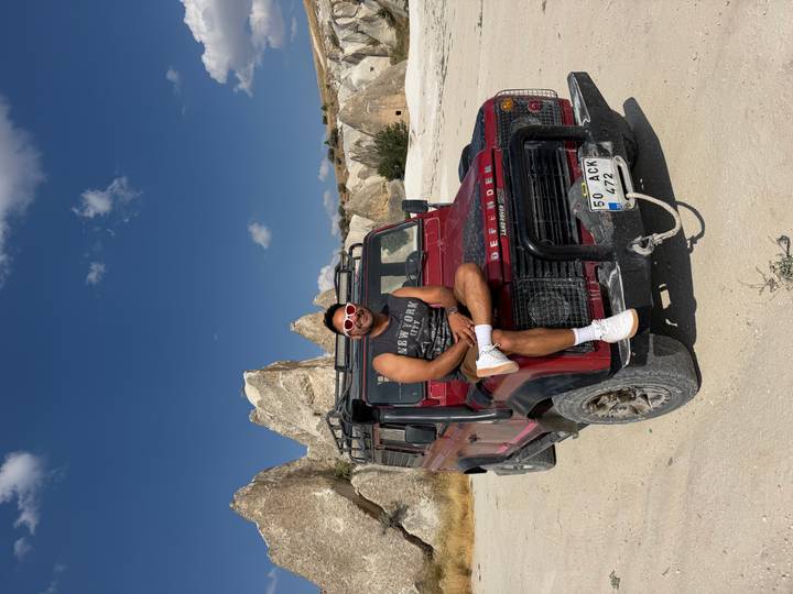 Young man in sunglasses posing on a red 4x4 jeep with Cappadocia rock formations behind