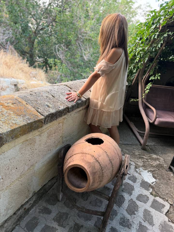 Woman in a light dress leaning on a stone terrace next to an old clay jar