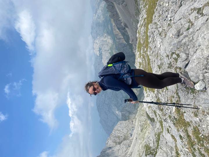 Female hiker with trekking poles standing on a rocky summit above cloud-draped mountains.