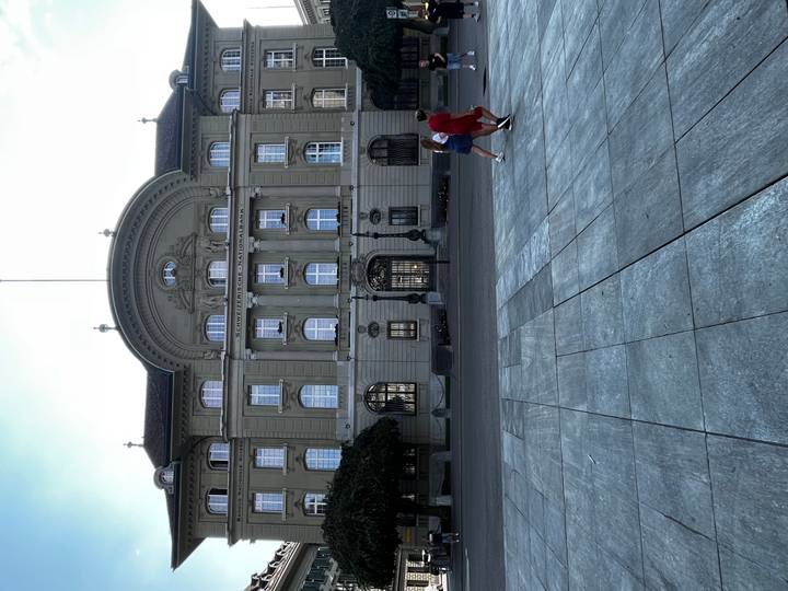 Grand historic government building with an ornate façade facing a wide stone plaza where two women walk across the foreground.