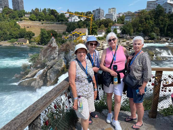 Four smiling women pose on a wooden viewing deck overlooking the powerful Rhine Falls cascading around a rocky islet.
