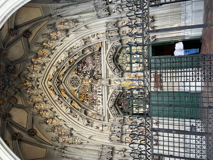 Highly detailed Gothic portal of a cathedral decorated with intricate stone carvings above green doors while two visitors enter.