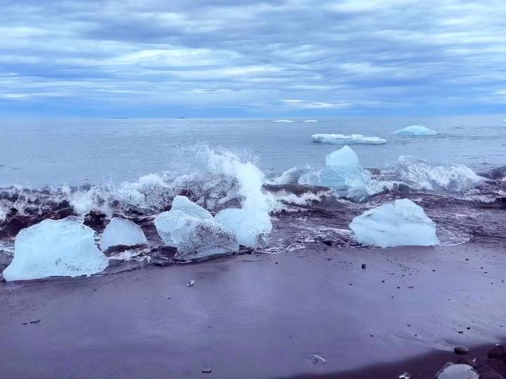 Waves crash over luminous blue ice chunks scattered on a black-sand beach under cloudy skies.