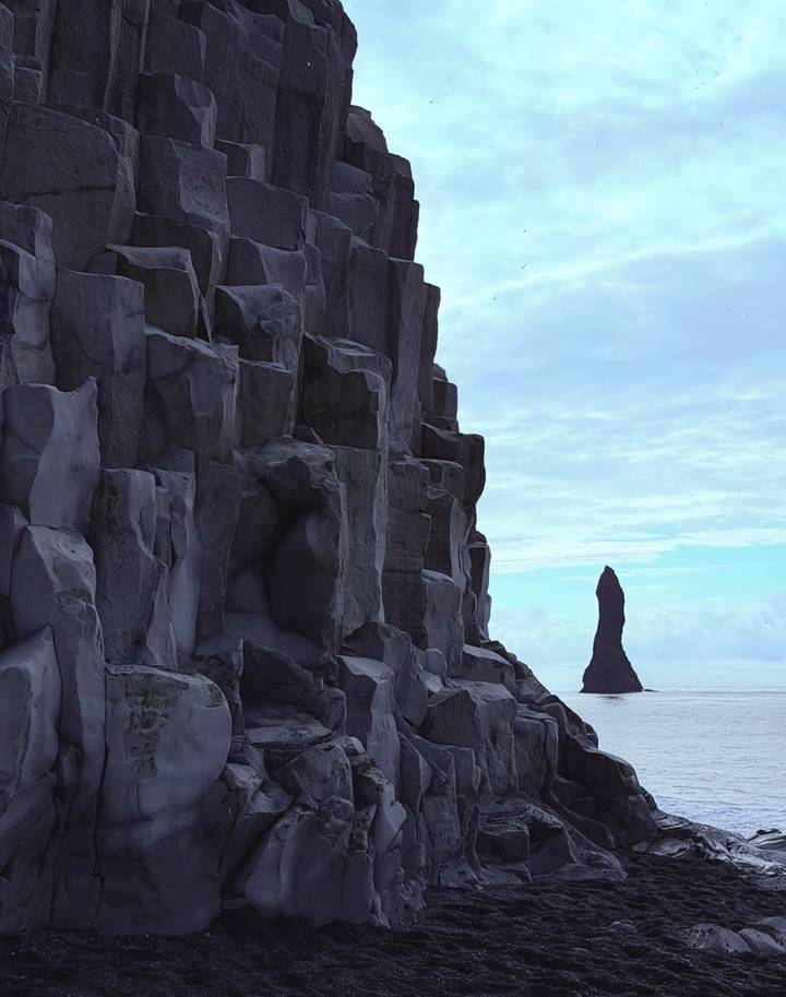 Tall basalt sea stack rises offshore beside dramatic hexagonal cliff columns under blue sky.