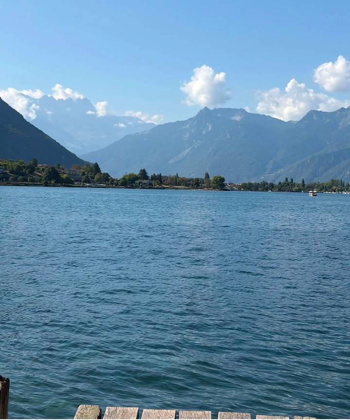 Calm blue lake framed by distant alpine mountains under a lightly clouded sky.