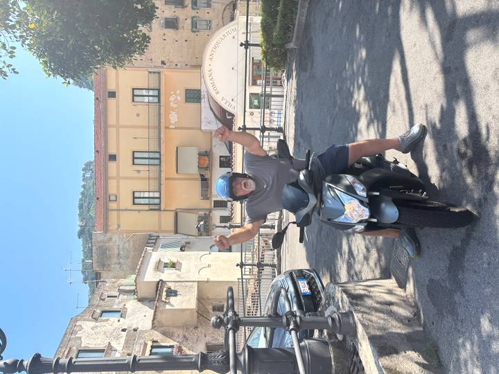 A smiling man wearing a helmet raises his hands triumphantly while sitting on a black scooter in a sunny Italian village street.
