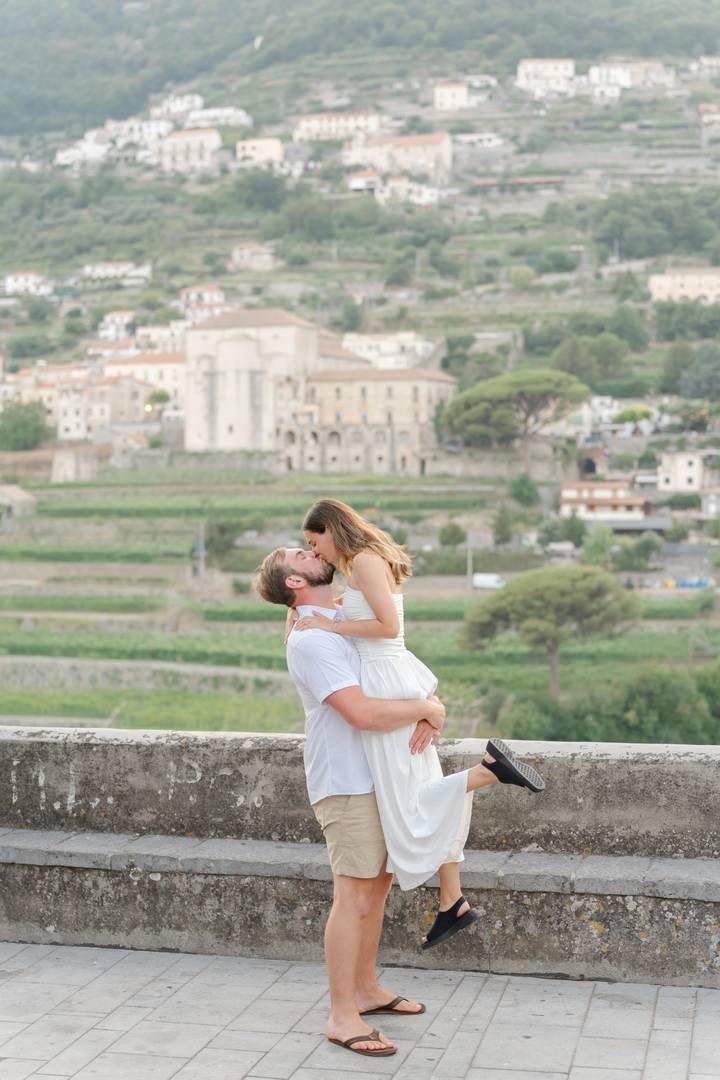 A romantic couple sharing a kiss while the man lifts the woman against a lush terraced hillside dotted with historic buildings.