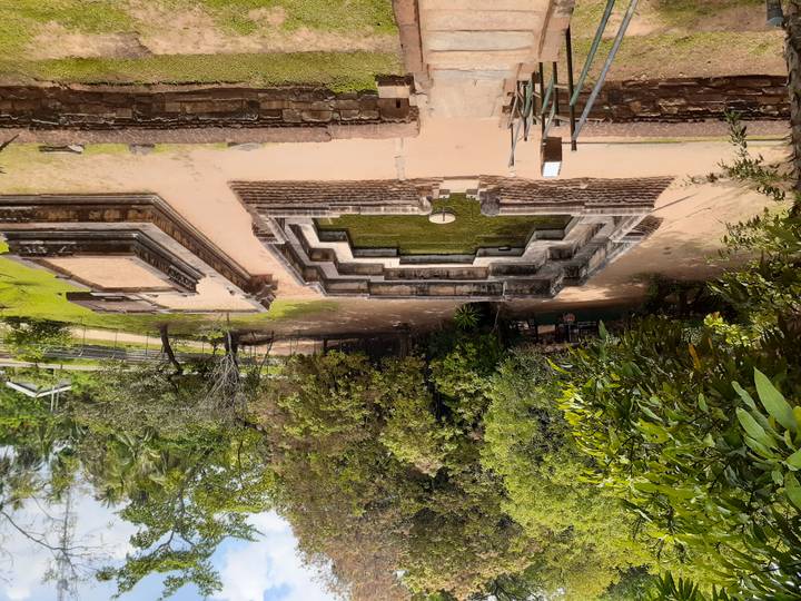 Ancient stepped stone pool surrounded by trees within an archaeological site, viewed from an elevated stairway.