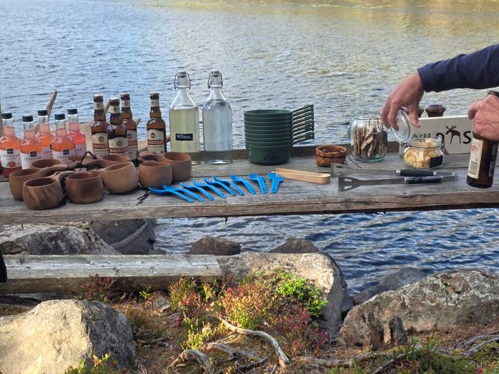 Rustic wooden table by lake set with drinks, bottles and snacks for outdoor picnic.