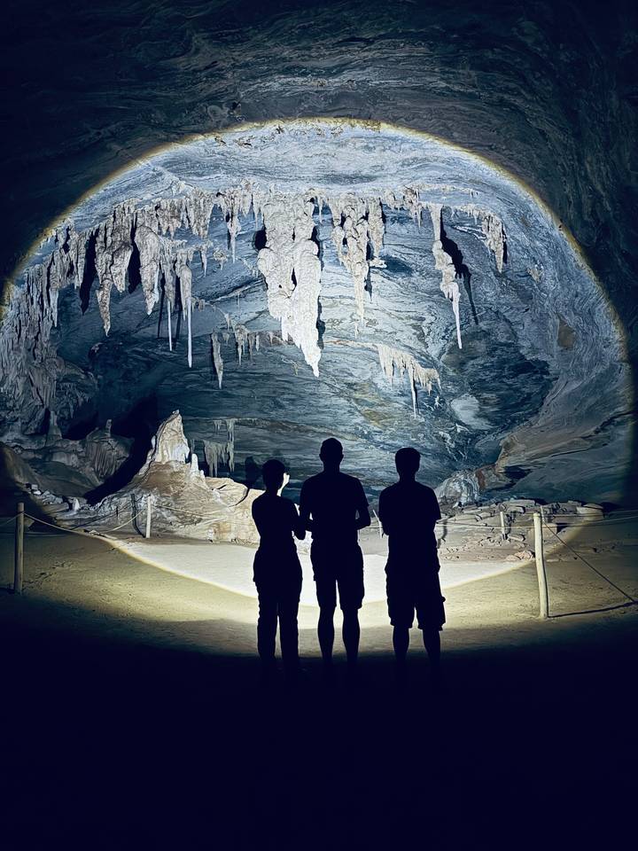 Silhouettes of three explorers facing an illuminated limestone cave interior with stalactites.