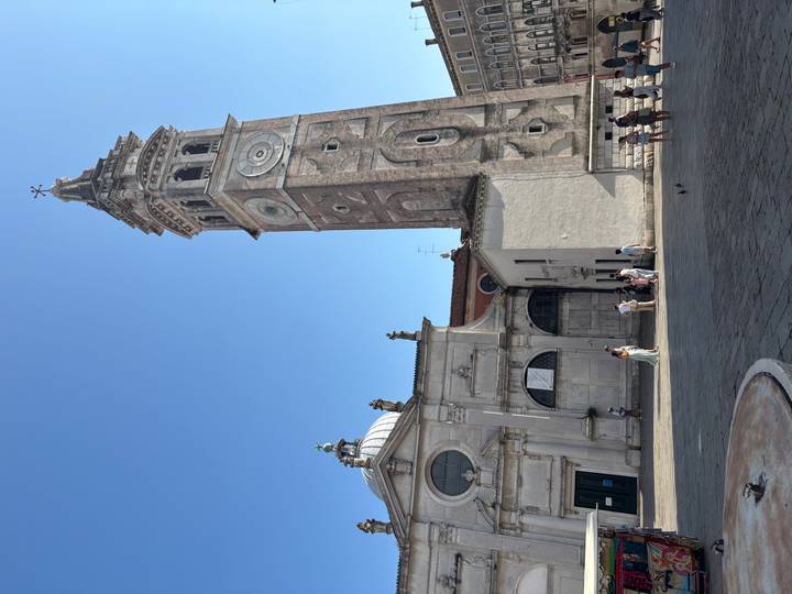 Tall stone clock tower and domed church overlook a sunny courtyard with scattered visitors.