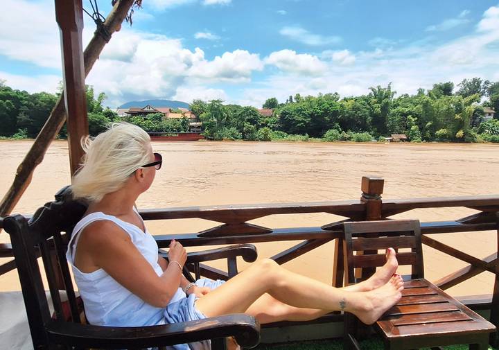 Woman relaxing on a wooden boat deck looking across a wide muddy river toward lush green banks.