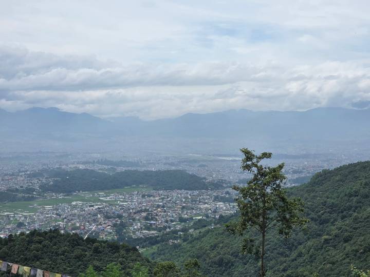 Wide view over Kathmandu Valley with cityscape, green hills and cloud-shrouded mountains beyond.