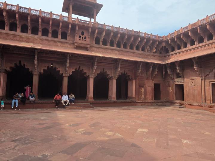 Spacious sandstone courtyard with ornate arches and visitors seated along the edge.