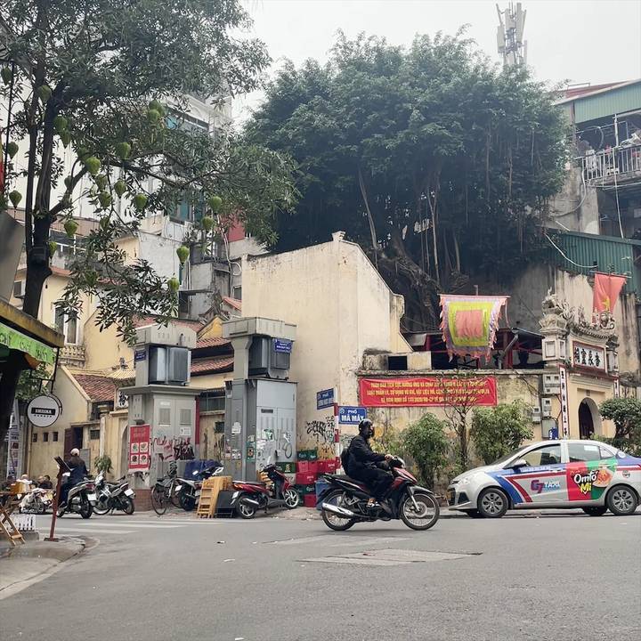 Bustling Vietnamese street corner with motorbikes, taxis, and colorful signage under leafy trees
