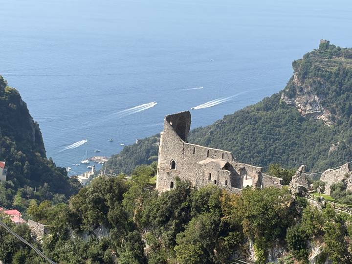 Ruined stone structure perched on a lush mountainside overlooking the Amalfi Coast and speeding boats