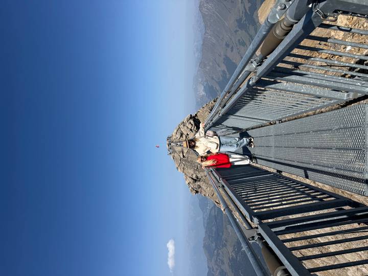 Two travelers standing on a high metal bridge walkway leading to a mountain peak with blue skies