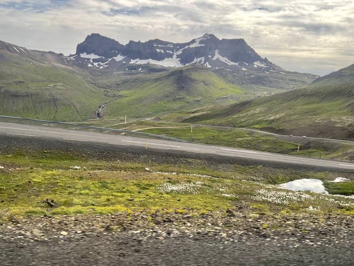 Sloping Icelandic mountain road cutting across lush valley with snow-specked peaks beyond.