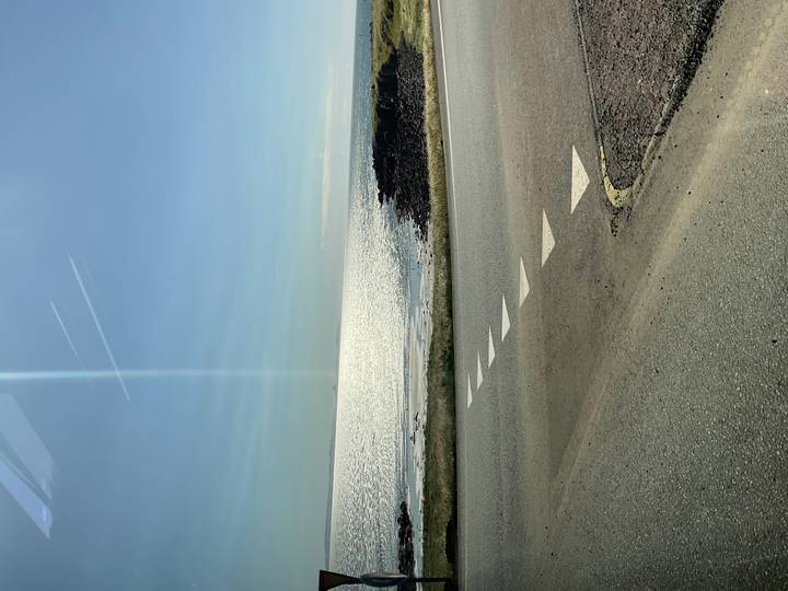 Glare-filled vertical shot of coastal road with sunlit sea and clear blue sky in Iceland.