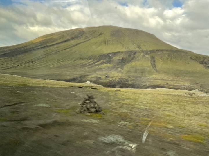Blurred foreground of rocky Icelandic plateau with conical hill under cloudy sky.
