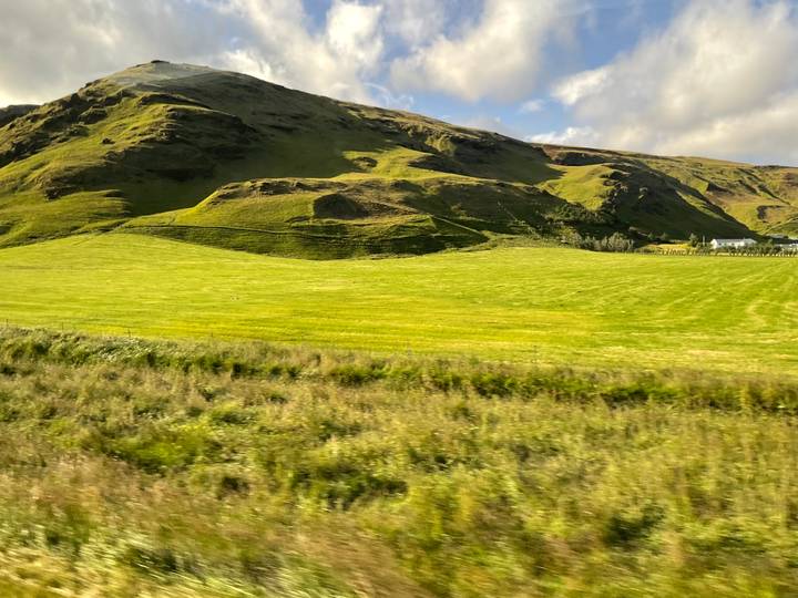 Grassy Icelandic meadow with rolling hills and farmhouse under partly sunny skies.