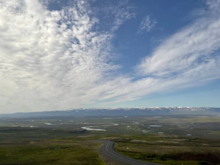 Wide view of Icelandic plain with braided rivers and distant snow-capped range beneath dramatic clouds.