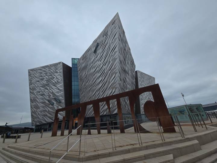 The angular silver facade of Titanic Belfast museum soars against a grey sky with a giant rusted sign in front.