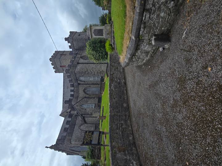 A grey-stone church with crenellated tower sits atop a damp gravel path after rainfall.