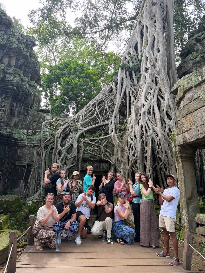 Tour group poses with playful hand gestures beneath massive tree roots enveloping ancient Angkor temple stonework.