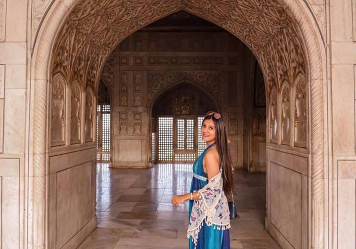 Woman in a blue dress looks back while walking through ornate marble corridors of Agra Fort.