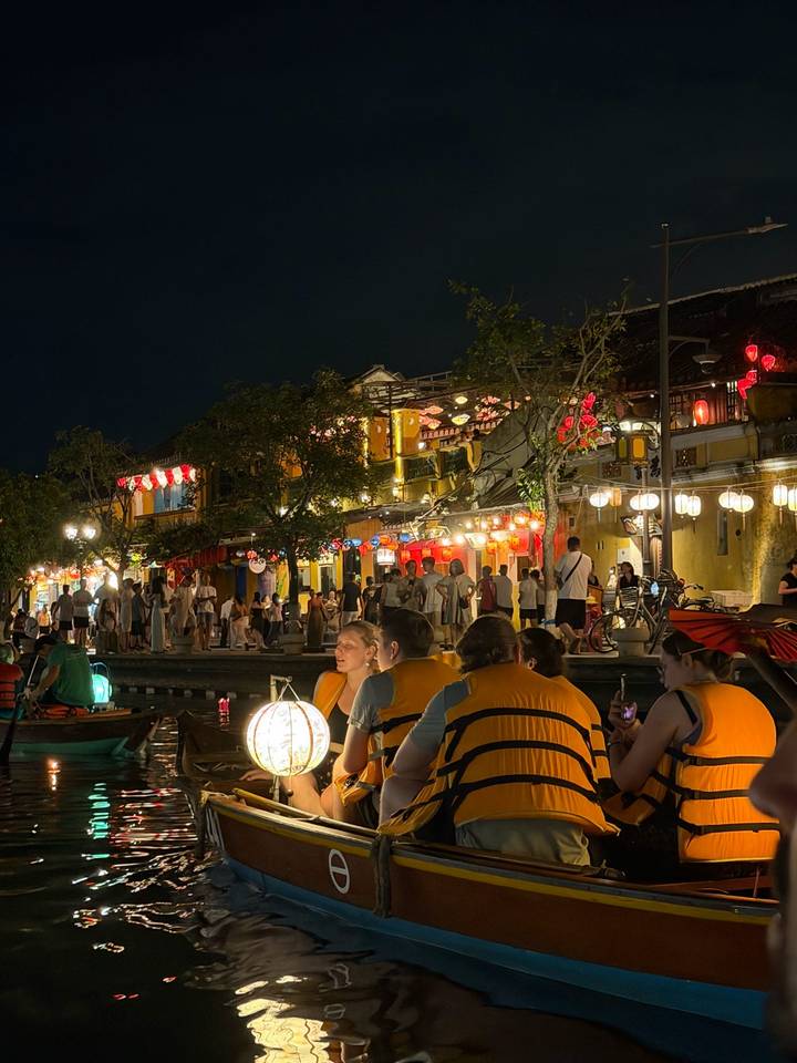 Nighttime riverside in Hoi An with illuminated lanterns, crowded street and tourists in boats wearing life vests.