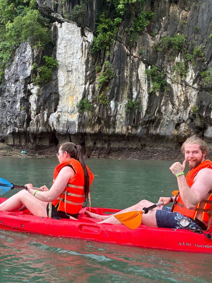Kayakers in orange life vests paddling near limestone cliffs on emerald water giving a thumbs up.