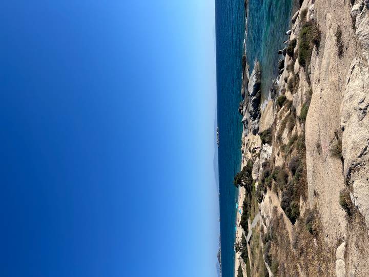 Rocky seaside landscape with clear Aegean water and visitors scattered along the shore.