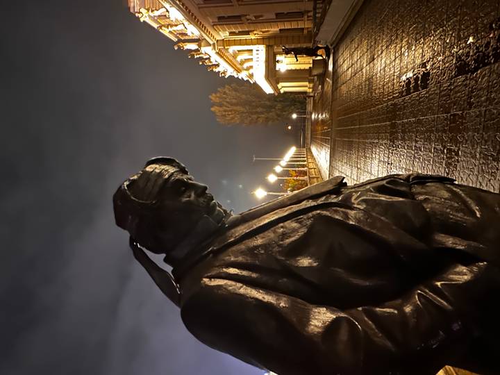 Bronze statue of a contemplative man at night on a rain-slick plaza with lit buildings behind.