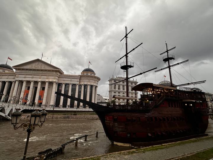Wooden themed restaurant ship moored on river beside neoclassical museum under stormy sky.