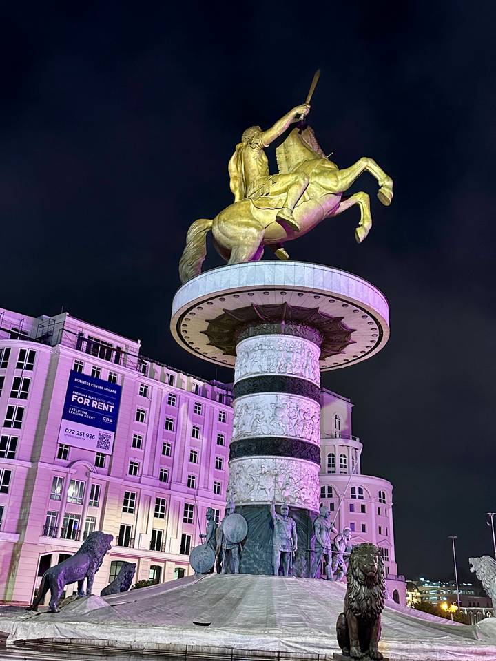 Illuminated Alexander the Great statue atop ornate column against night sky with nearby buildings lit purple.