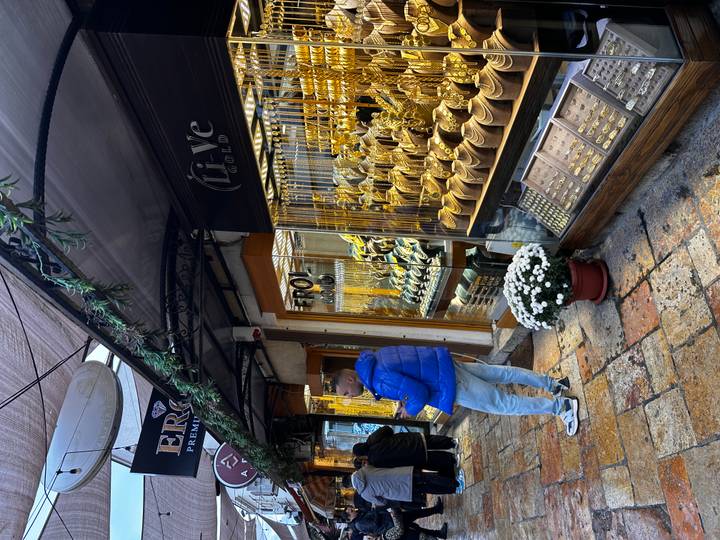 A narrow bazaar lane with brightly lit gold jewelry shops and a young man in a blue jacket browsing.