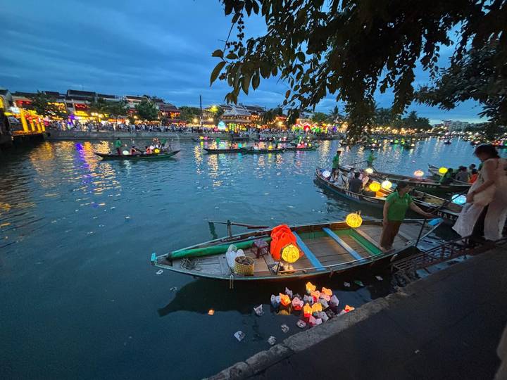 Evening river scene with lantern-lit boats and crowds along illuminated waterfront promenade.
