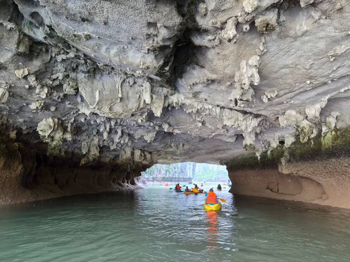 Adventurers paddling kayaks through a dramatic cave tunnel with stalactites and turquoise water.