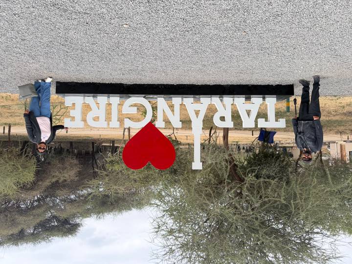 Two visitors posing beside the large 'I ♥ Tarangire' park sign in a dry woodland setting.