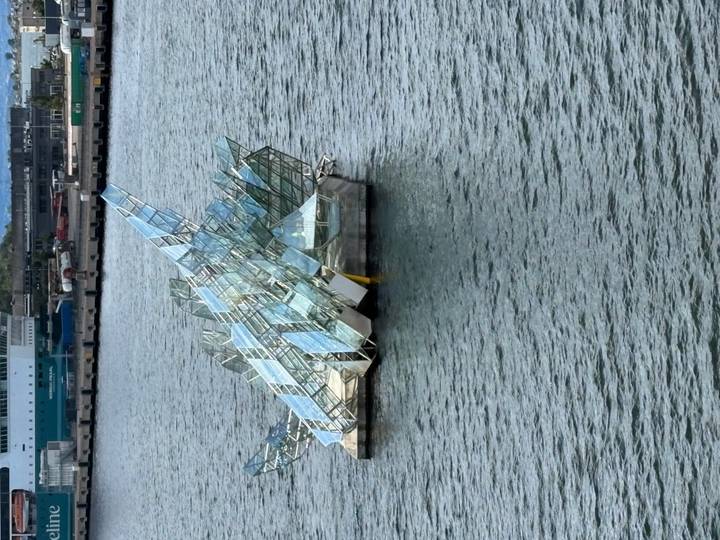 Floating glass sculpture on calm harbor water with a ferry terminal in the background.